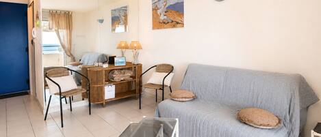 Una sala de estar con un sofá, sillas, una mesa de vidrio y un espejo que refleja una cama y cortinas.