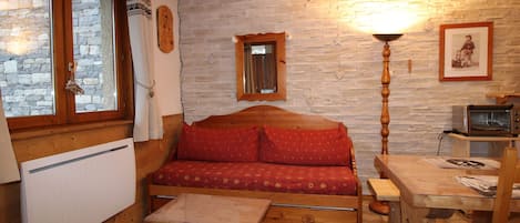 Una habitación acogedora con un sofá de madera, una mesita, una lámpara, un ventanal con cortinas, una pared de piedra y un cuadro enmarcado.