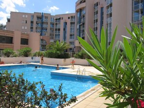 Piscina com pessoas ao redor, cercada por um edifício com varandas e vegetação.