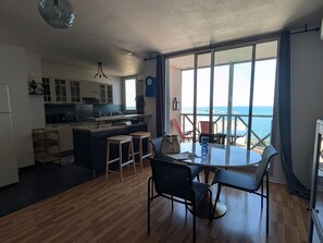 Une cuisine moderne dotée d’un îlot central, d’un coin repas avec une table et des chaises, et d’une grande fenêtre offrant une vue sur la mer.