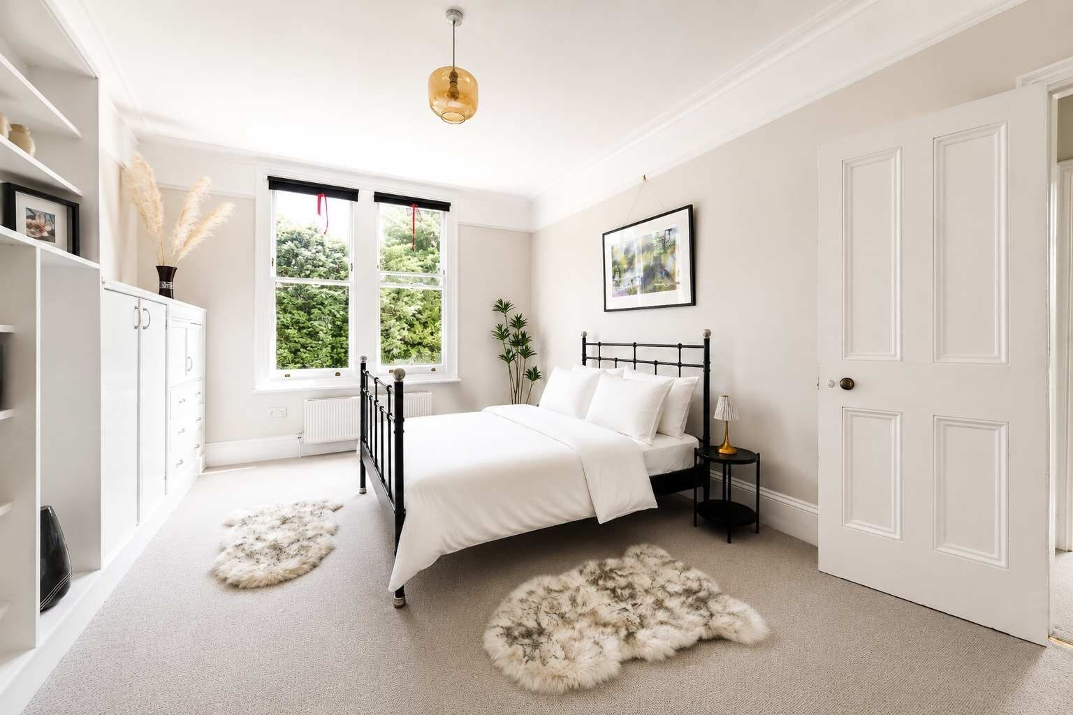 A bedroom with a bed, a nightstand, a lamp, a door, a window, and a radiator.