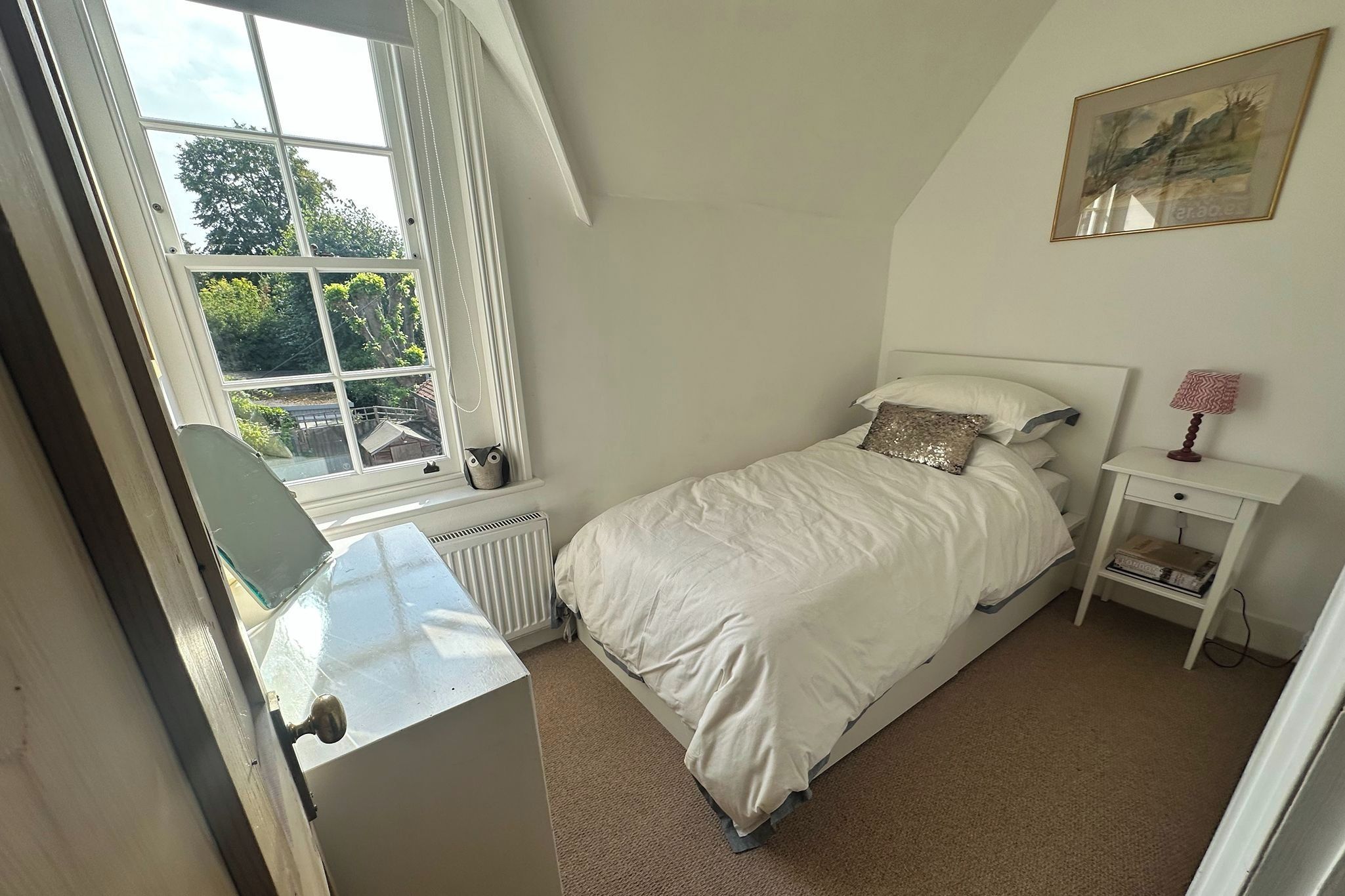 Quarto com cama, mesa de cabeceira, radiador e janela com cortinas.