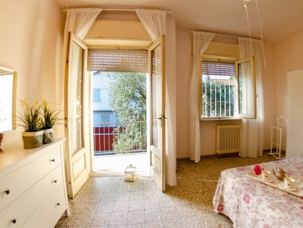 A bedroom with a bed, a dresser, a window with curtains, a radiator, and a balcony with a view of buildings and trees. - Angelina by Interhome (Lido di Camaiore)