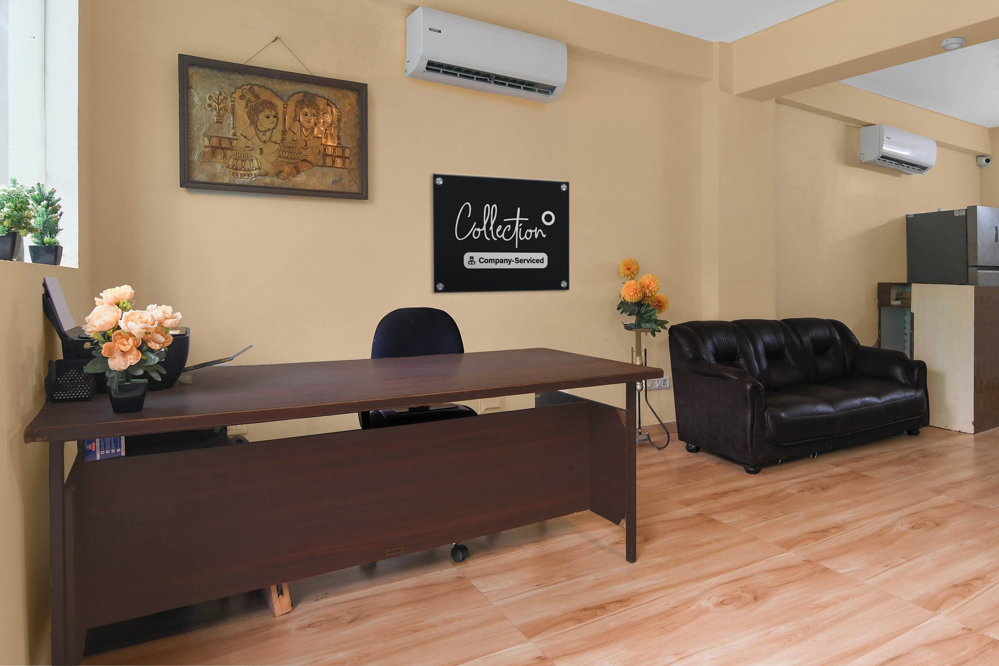 A furnished room with a wooden desk, a black leather couch, a framed artwork, and a sign indicating the room is company-serviced.
