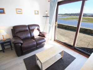 A living room with a brown leather sofa, a small wooden coffee table, a lamp, and a view of a river through a glass door. - Aldena 12a South Snowdon Wharf (Porthmadog)