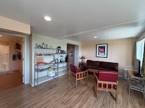 Una sala de estar con un sofá marrón, dos sillas de madera, una mesa de centro, un televisor y una estantería con utensilios de cocina.