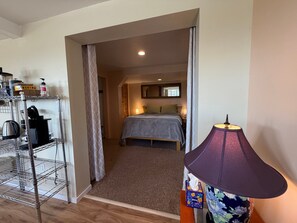 Habitación de hotel con cama, mesita de noche, lámpara, repisa con artículos de aseo y puerta que da a otra habitación.