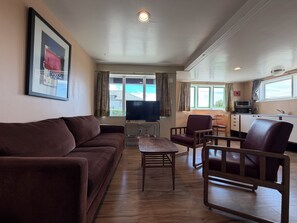 A living room with a sofa, a coffee table, and a TV. There is a kitchen area with a microwave and a sink.