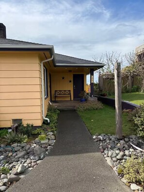 A residential property with a paved driveway leading to a front door, a small porch with a bench, and a garden with various plants and a wooden fence.
