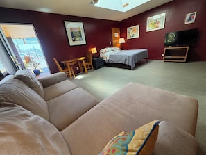 A room with a sofa, a bed, a desk, and a TV. There are paintings on the walls and a window with a view.