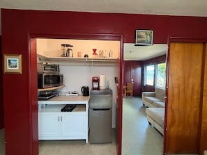A compact kitchen with a microwave, coffee maker, and refrigerator. There is a dining area with a table and chairs, and a sofa in the adjacent living room.