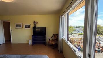 Un dormitorio con cama, televisor sobre un mueble, una silla y ventana con vistas al exterior.