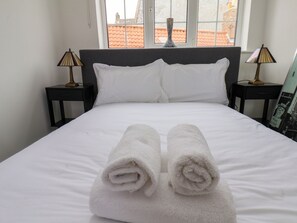 A neatly made bed with white linens and two rolled towels in the center, flanked by two bedside tables with lamps, in a room with a window overlooking a red-tiled roof. - Og's House (Whitby)