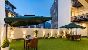 Un espacio exterior bien iluminado para comer, con mesas y sillas, rodeado de plantas en macetas y un césped.