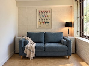 Ein Zimmer mit einem blauen Sofa, einem gerahmten Poster an der Wand, einer Lampe und einem Fenster mit Blick.