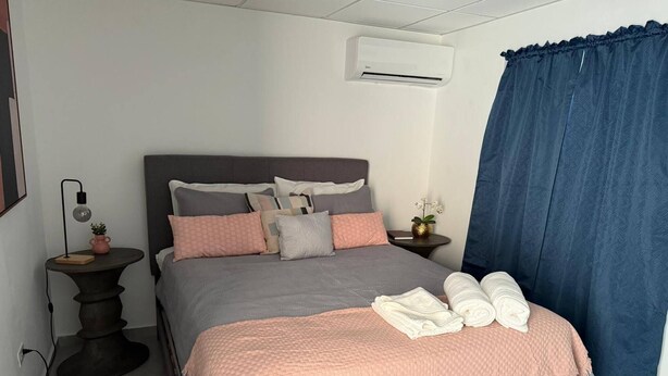 Ein ordentlich eingerichtetes Schlafzimmer mit Bett, Kissen, Nachttisch mit Lampe, einer kleinen Pflanze und einem Fenster mit Vorhängen.