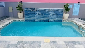 Ein Schwimmbecken mit einer Fliesenleiste, umgeben von einer gefliesten Terrasse und Pflanzen in Töpfen auf beiden Seiten.