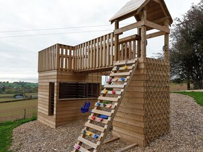 A wooden play structure with a slide and climbing wall, surrounded by a gravel area and trees. - Providence Cottage (Monmouth)