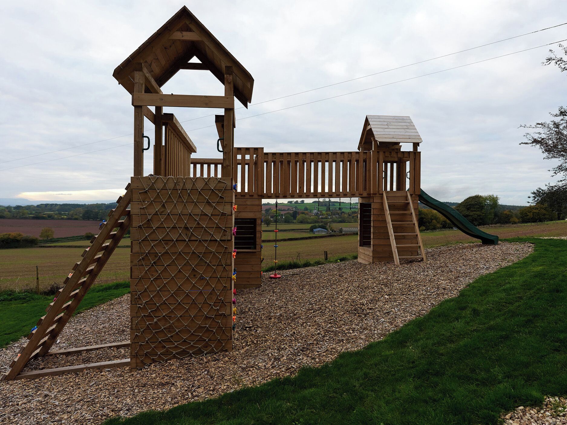 A wooden playground structure with a slide and climbing elements, surrounded by gravel and grass.