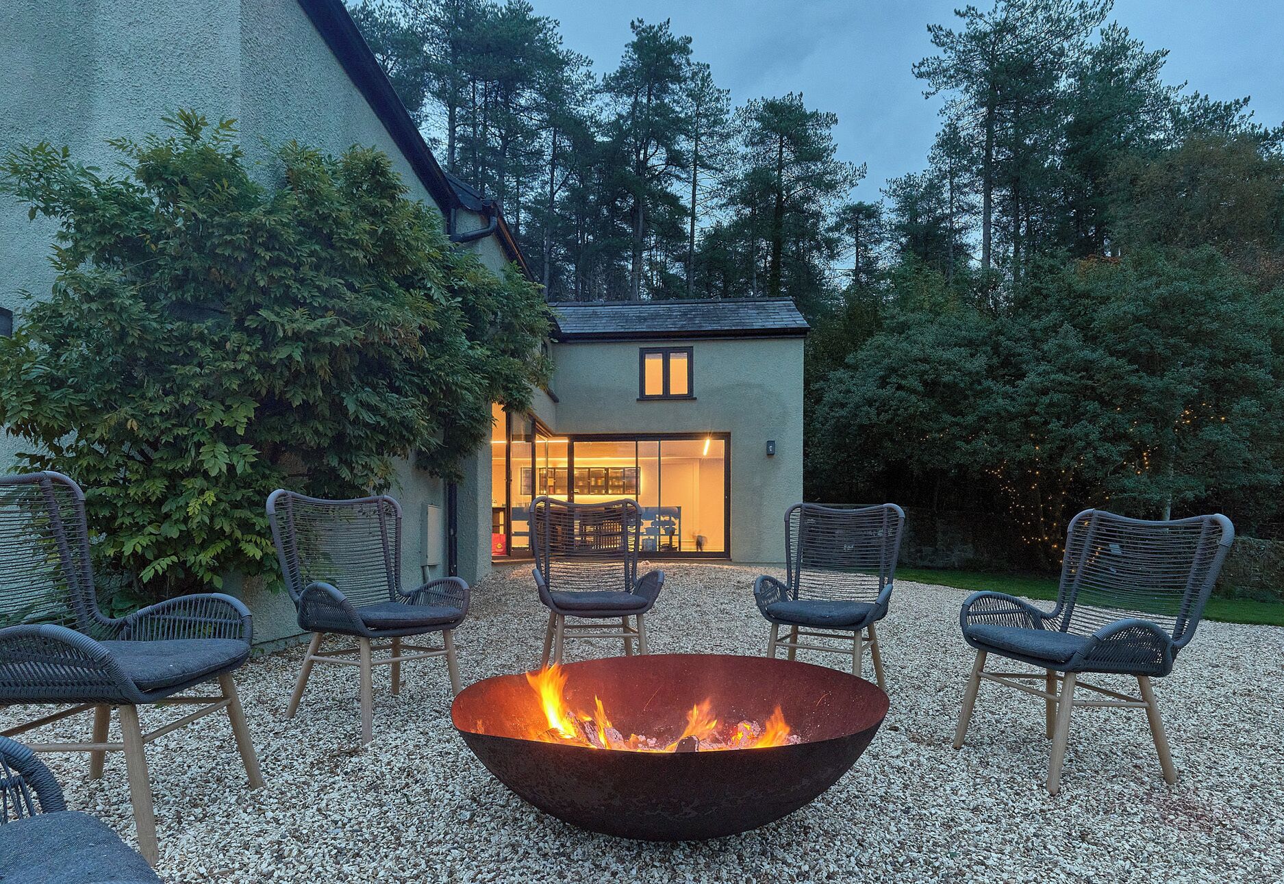 A patio with a fire pit surrounded by chairs, a building with a glass door, and a gravel ground.