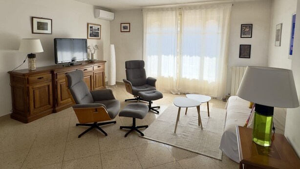 Een woonkamer met een televisie, een houten kast, twee stoelen, een klein tafeltje en een bank.