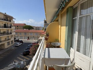 A balcony with a checkered table, potted plants, and a view of a street with parked cars and buildings. - (026) Le côté Remparts (Antibes)