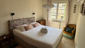 Een slaapkamer met een bed, nachtkastjes, een stoel, een raam met gordijnen, en een luchtverkoeler aan de muur.