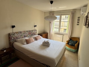 A bedroom with a bed, bedside tables, a chair, a window with curtains, and a wall-mounted air conditioner. - (026) Le côté Remparts (Antibes)
