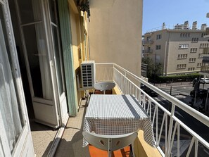 A balcony with a checkered table and chairs, an air conditioning unit, and a view of a cityscape with buildings. - (026) Le côté Remparts (Antibes)