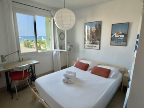 A hotel room with a bed, a desk, a chair, a fan, and a view of the sea. - (026) Le côté Remparts (Antibes)