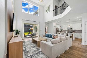 A spacious living area with a large sofa, a coffee table, and a TV mounted on the wall. There is a sliding glass door leading to an outdoor patio area. - Brand New 5BR 5BA Villa w Pool Spa Game Room 7800 (Kissimmee)