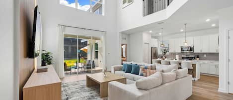 A spacious living room with a large sofa, a coffee table, and a TV mounted on the wall. There is a staircase leading to an upper level.