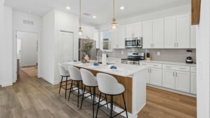 A modern kitchen with white cabinetry, a central island with bar stools, stainless steel appliances, and pendant lighting. - Brand New 5BR 5BA Villa w Pool Spa Game Room 7800 (Kissimmee)