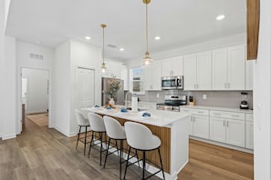 A modern kitchen with white cabinetry, a central island with bar stools, stainless steel appliances, and pendant lighting. - Brand New 5BR 5BA Villa w Pool Spa Game Room 7800 (Kissimmee)