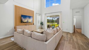 A spacious living room with a large sectional sofa, a wooden accent wall, and a large window offering a view of the outdoors. - Brand New 5BR 5BA Villa w Pool Spa Game Room 7800 (Kissimmee)