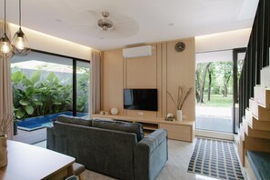Ein modernes Wohnzimmer mit Flachbildfernseher, grauen Sofas, einem Esstisch und einem großen Fenster mit Blick auf Grünflächen.