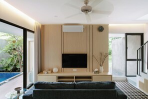 Ein modernes Wohnzimmer mit Flachbildfernseher, Holzwandverkleidung, Deckenventilator und Blick auf einen Swimmingpool.