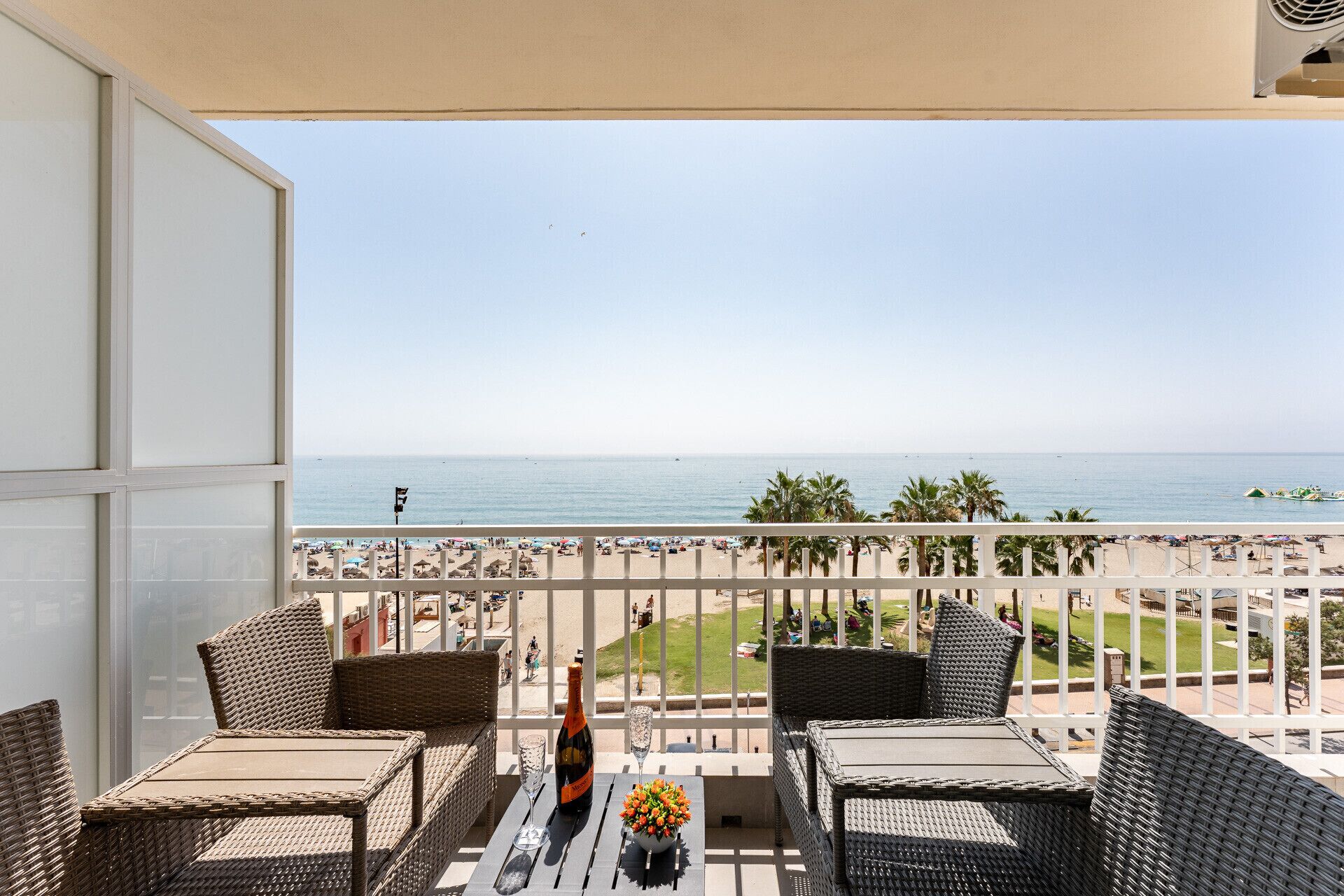 A balcony with wicker furniture, a bottle of champagne, and a glass. The balcony overlooks a beach with palm trees and the sea.
