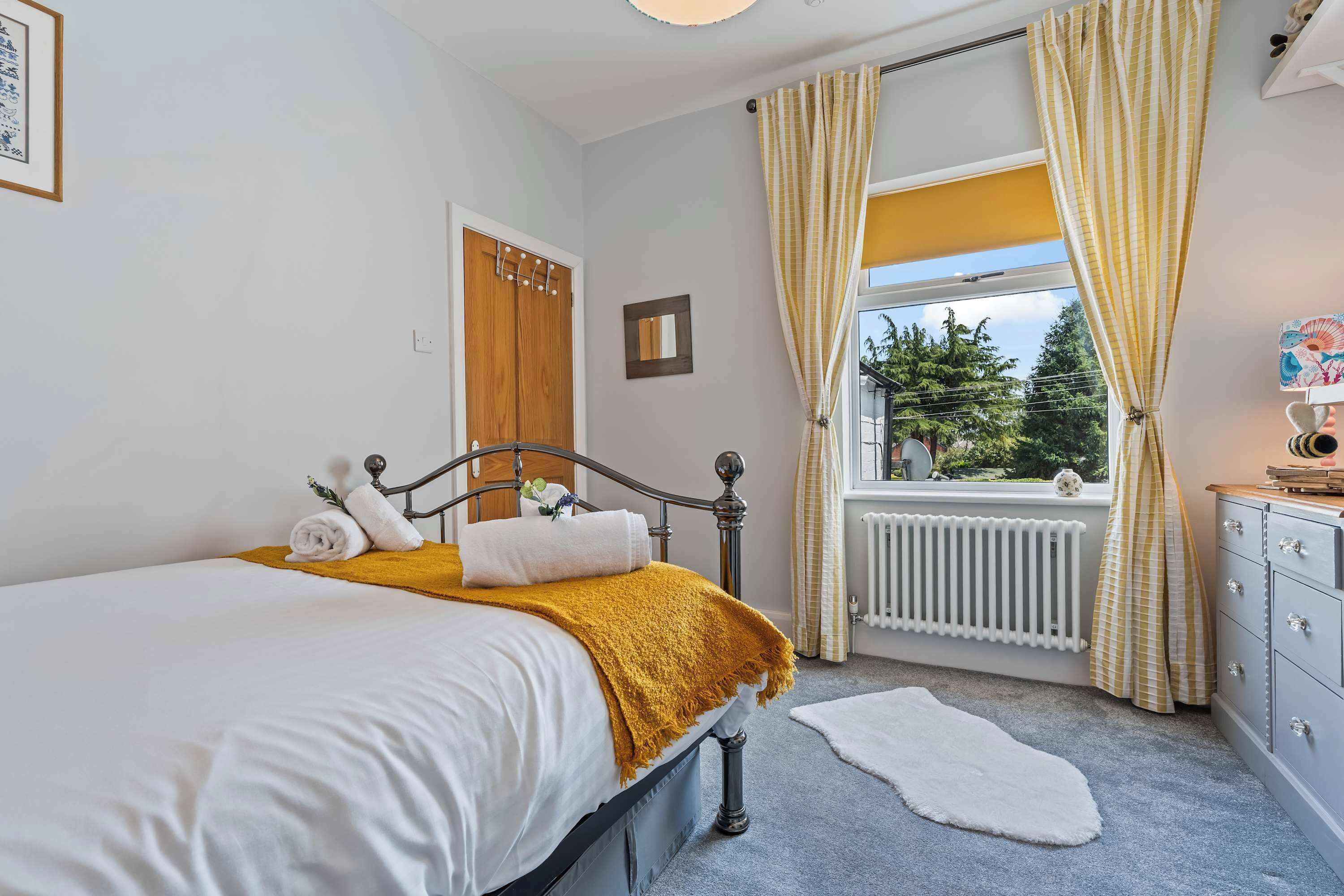 A bedroom with a bed, a dresser, a window with curtains, a radiator, and a small rug.