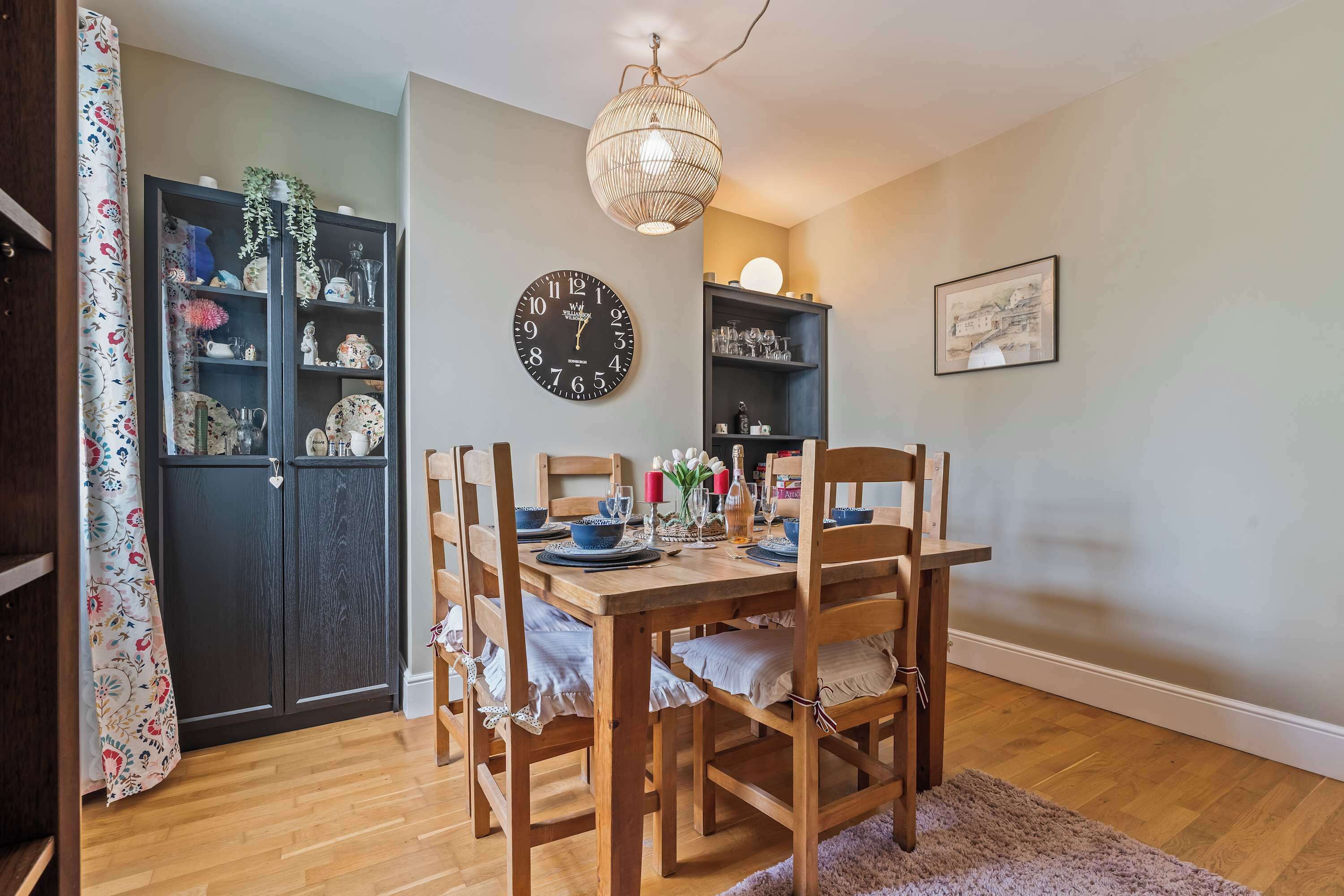 A dining area with a wooden table set for four, surrounded by chairs. A wall clock and shelves with various items are visible.