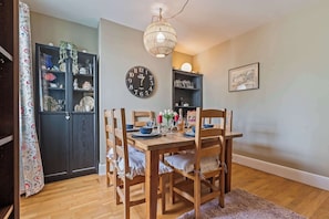 A dining area with a wooden table set for four, surrounded by chairs. A wall clock and shelves with various items are visible. - Charming 2-bed Home Away From Home in Nantwich (Nantwich)