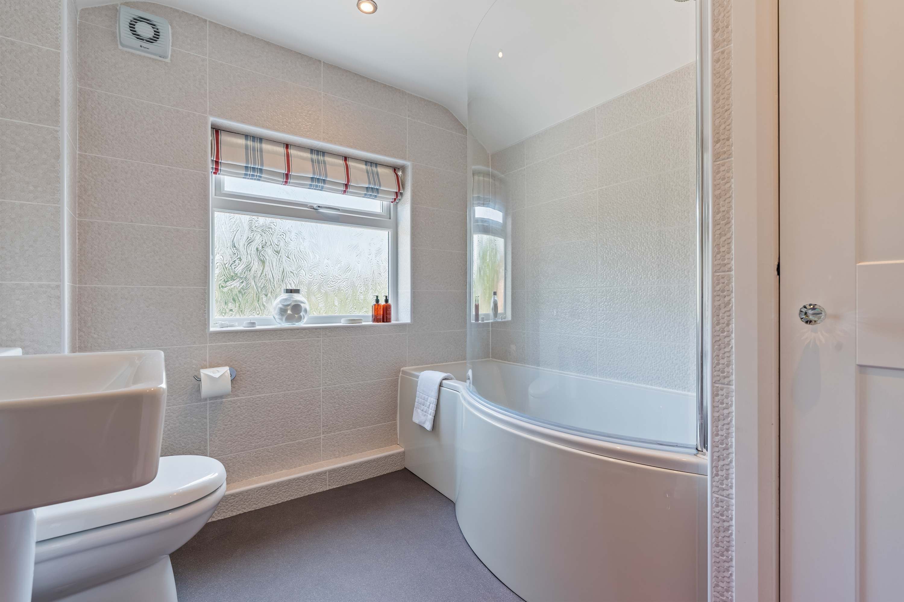 A modern bathroom with a toilet, a window with a striped blind, and a bathtub.
