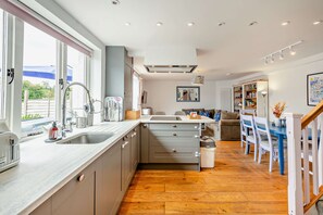 A modern kitchen with a central island, built-in appliances, and a dining area with a blue table and chairs. - Host Stay The Hidden House (Dawlish)