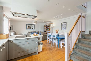 A modern kitchen with grey cabinets, a dining area with blue chairs, and a staircase with wooden railings. - Host Stay The Hidden House (Dawlish)