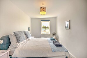 Un dormitorio con cama, almohadas, mesita de noche, ventana, radiador, un cuadro en la pared y una lámpara en el techo.