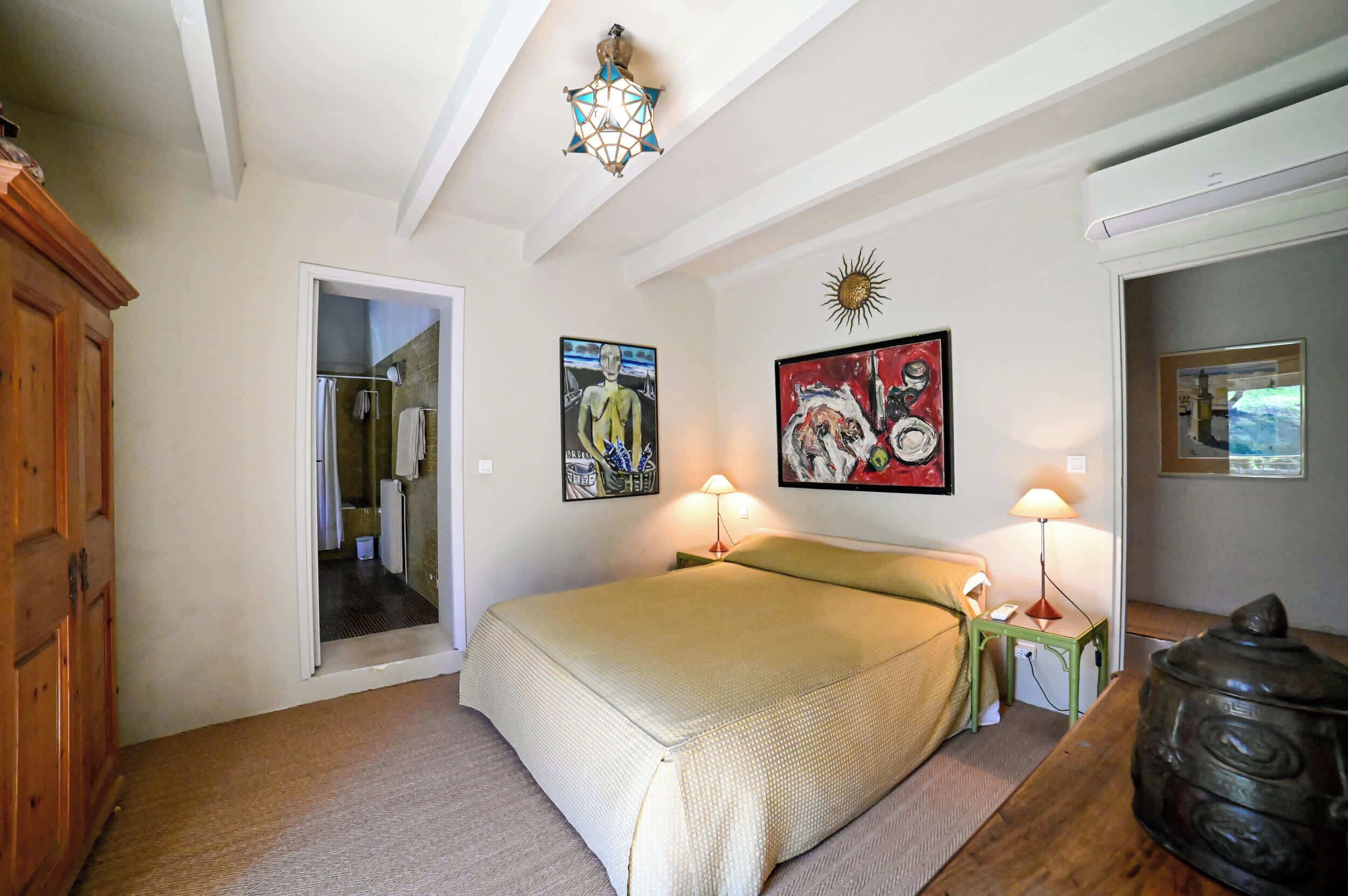 A bedroom with a bed, two bedside lamps, a wooden cabinet, a painting, and a sun decoration on the wall.