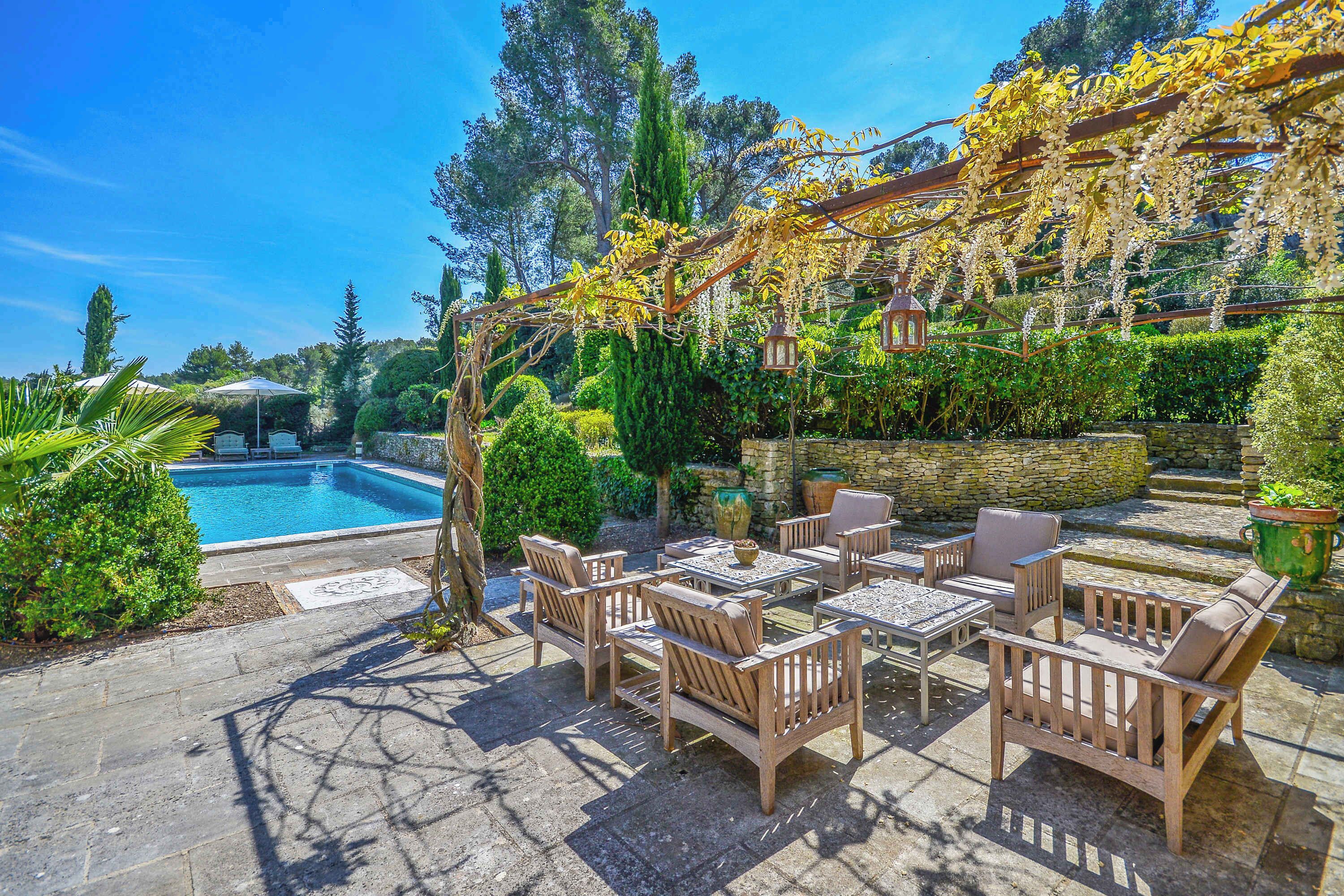 An outdoor patio with a pool, wooden furniture, and a pergola covered with flowering vines.