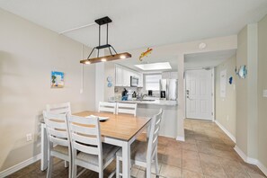 A dining area with a wooden table and white chairs, a kitchen with white cabinets, and a white door. - Sunswept 102 by Liquid Life (Orange Beach)