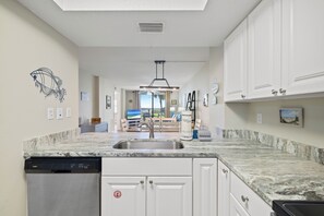 A kitchen with a built-in dishwasher, white cabinetry, a marble countertop, and a view of the living area through an open door. - Sunswept 102 by Liquid Life (Orange Beach)