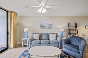 A living room with a grey sofa, two side tables with lamps, a ceiling fan, a painting of a beach, and a bookshelf. - Sunswept 102 by Liquid Life (Orange Beach)